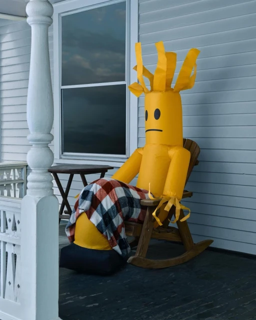 A yellow inflatable tube man sits in a rocking chair on a porch with a plaid blanket draped over it, creating a humorous and whimsical scene.